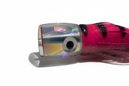 A close-up of a fishing lure with a clear, holographic head, a large black-and-white eye, and a bright pink skirt with black stripes and glitter, resting on a beige fabric surface.