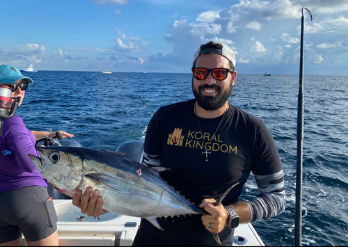 A man on a boat wearing a black Koral Kingdom long-sleeve shirt holds a large tuna fish, with another person partially visible on the left and an ocean background under a blue sky.